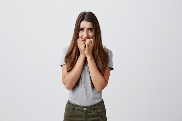 Hermosa chica infeliz estudiante caucásica con cabello largo y oscuro en camisa gris y jeans royendo los dedos con expresión asustada, asustada con sonidos extraños