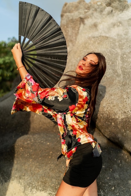 Hermosa chica japonesa en kimono elegante con gran abanico y maquillaje profesional posando sobre rocas