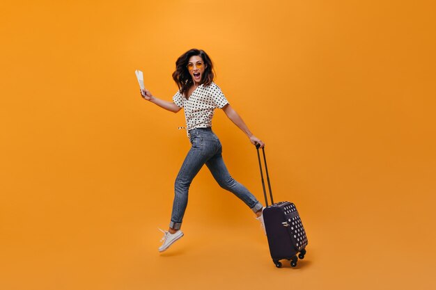 Hermosa chica en jeans está saltando sobre fondo naranja. Retrato de cuerpo entero de mujer con billetes y maleta