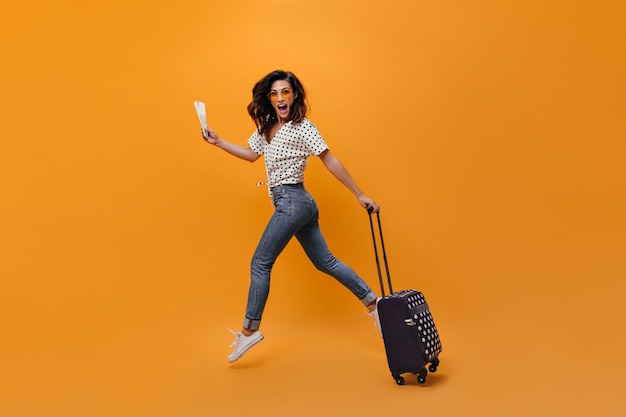 Hermosa chica en jeans está saltando sobre fondo naranja. Retrato de cuerpo entero de mujer con billetes y maleta