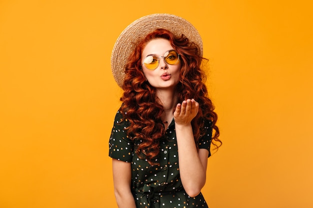 Hermosa chica de jengibre enviando beso de aire sobre fondo amarillo. Disparo de estudio de mujer joven rizada en gafas de sol y sombrero de paja.