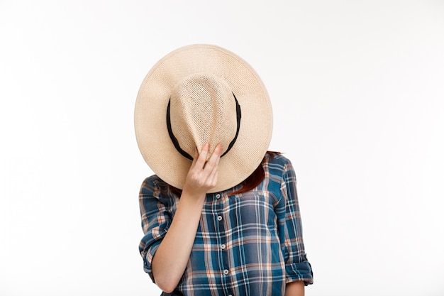 Hermosa chica jengibre escondiendo su rostro con sombrero sobre pared blanca.