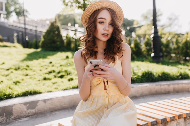 Hermosa chica de jengibre posando mientras envía mensajes de texto por la mañana. Disparo al aire libre de fascinante joven con sombrero sentado en el parque y disfrutando del clima de verano.