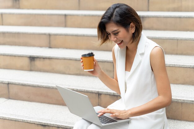 Hermosa chica linda sonriendo en ropa de mujer de negocios usando la computadora portátil en la ciudad urbana