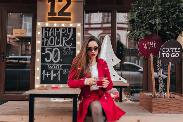 Hermosa chica lleva gafas de sol y falda roja sentada en la cafetería al aire libre