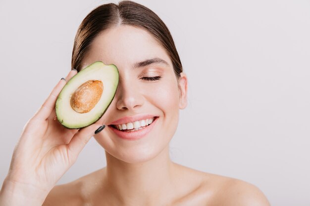 Hermosa chica sin maquillaje posando con los ojos cerrados en la pared blanca. Modelo sonriente cubre la cara con aguacate saludable.