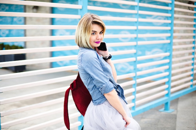 Hermosa chica medio vuelta con labios rosados brillantes sosteniendo un teléfono inteligente con rayas azules y blancas en el fondo.