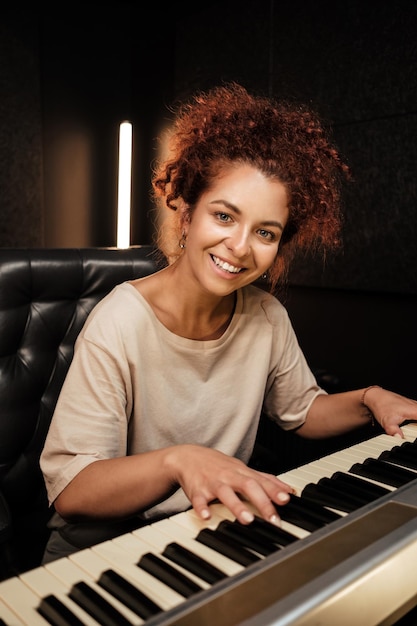 Hermosa chica mirando felizmente a la cámara tocando el piano eléctrico en el estudio. Músico atractivo trabajando en estudio de grabación de sonido