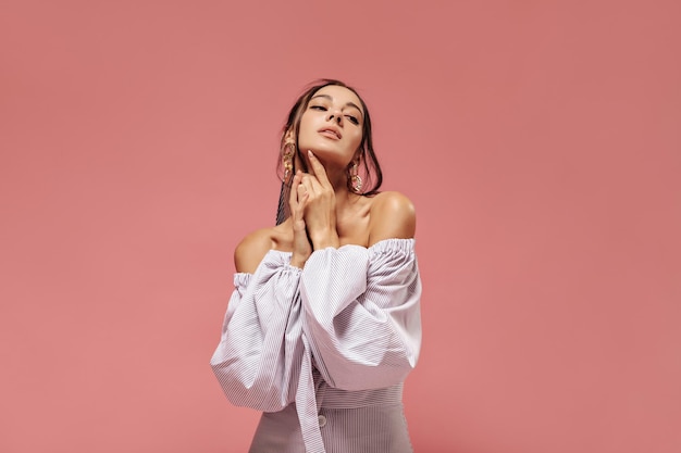 Hermosa chica moderna bronceada con cabello moreno en aretes y ropa ligera con estilo posando sobre fondo rosa aislado