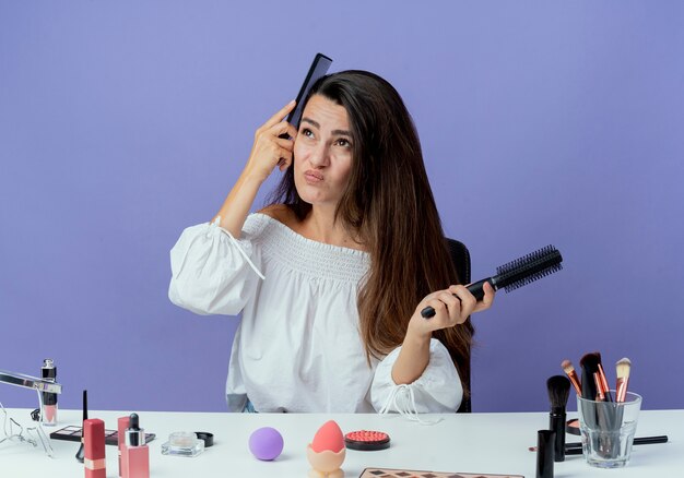 Hermosa chica molesta se sienta a la mesa con herramientas de maquillaje tiene peines mirando hacia arriba aislado en la pared púrpura