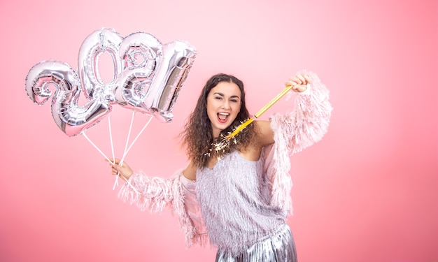 Hermosa chica morena alegre vestida de fiesta con el pelo rizado posando sobre un fondo de estudio rosa con globos plateados para el concepto de año nuevo