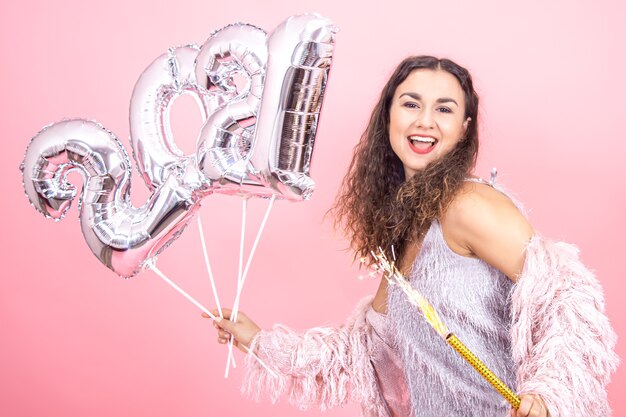 Hermosa chica morena alegre vestida de forma festiva con el pelo rizado sobre un fondo de estudio rosa posando con una vela de fuegos artificiales en la mano y globos plateados para el concepto de año nuevo