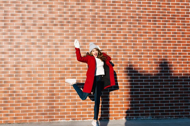 Hermosa chica morena con cabello largo divirtiéndose en la pared exterior. Viste abrigo rojo, guantes blancos y gorro de punto.