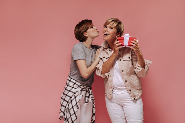 Hermosa chica morena en camisa y pantalón besando a su abuela en la mejilla sobre fondo rosa. Mujer rubia posando con caja de regalo roja y dama fresca.