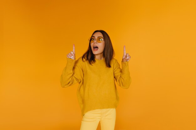 Hermosa chica morena con gafas posando en la pared amarilla. Mujer europea de moda que expresa asombro.