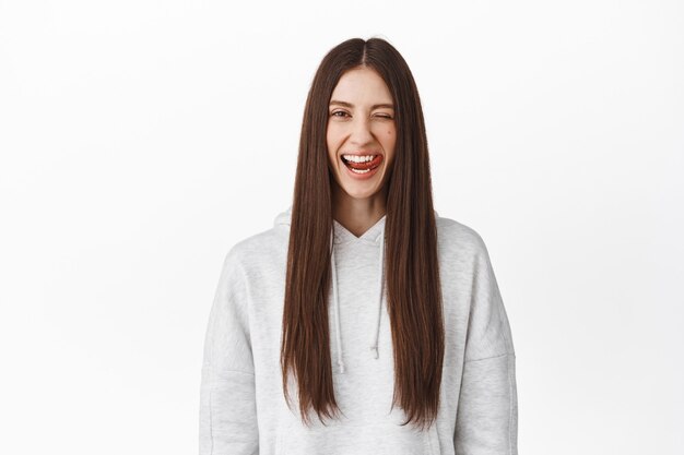 Hermosa chica morena con el pelo largo y liso, guiñando un ojo y mostrando la lengua, sonriendo dientes perfectos blancos, de pie contra la pared del estudio