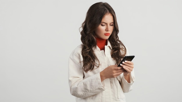 Hermosa chica morena seria atentamente usando smartphone sobre fondo blanco.