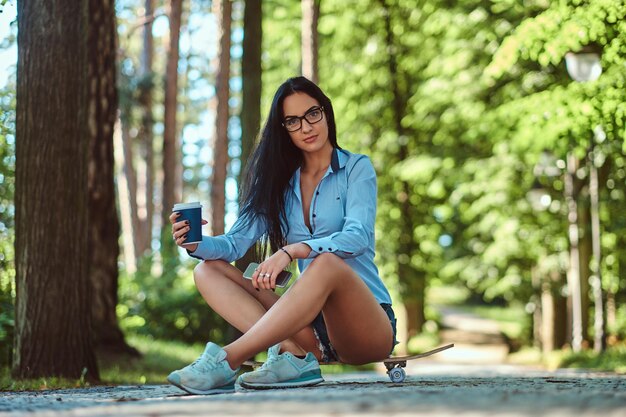 Hermosa chica morena sexy con gafas con una camisa y pantalones cortos sentada en una patineta y sostiene una taza de café para llevar y un teléfono inteligente en un parque.