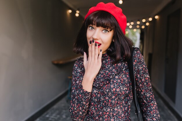 Hermosa chica morena con sombrero rojo con corte de pelo corto y poses de manicura oscura, con la mano tapándose la boca. Hermosa mujer joven en vestido estampado floral de pie en el bar con expresión de asombro