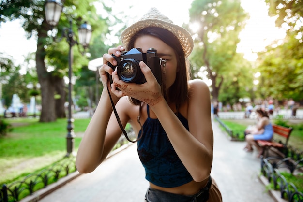 Hermosa chica morena con sombrero sonriendo, tomando fotos en el parque.