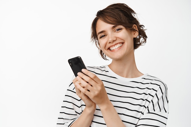 Hermosa chica morena con teléfono inteligente celular, sosteniendo el teléfono móvil y sonriendo feliz. Concepto de tecnología