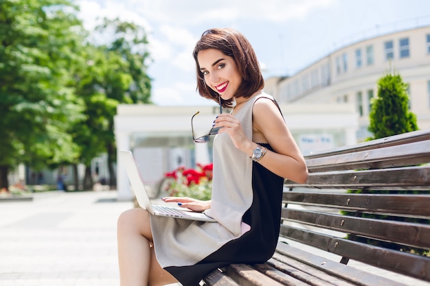 Una hermosa chica morena vestida de gris y negro está sentada en el banco de la ciudad. Ella tiene una computadora portátil en las rodillas y sonríe amigablemente a la cámara.