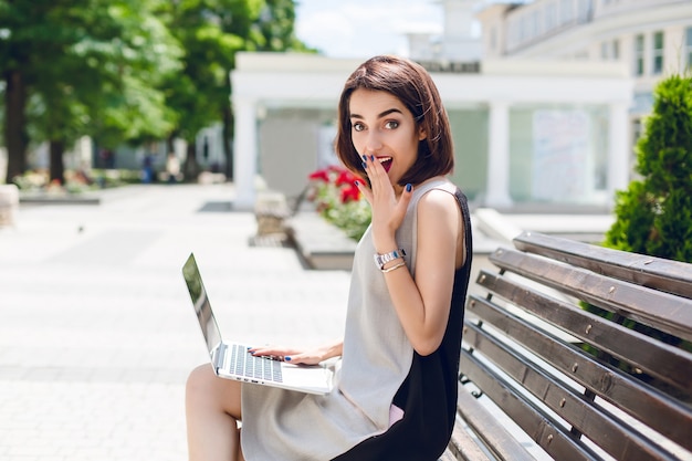 Una hermosa chica morena vestida de gris y negro está sentada en el banco de la ciudad. Ella tiene una computadora portátil en las rodillas y se ve sorprendida y divertida.