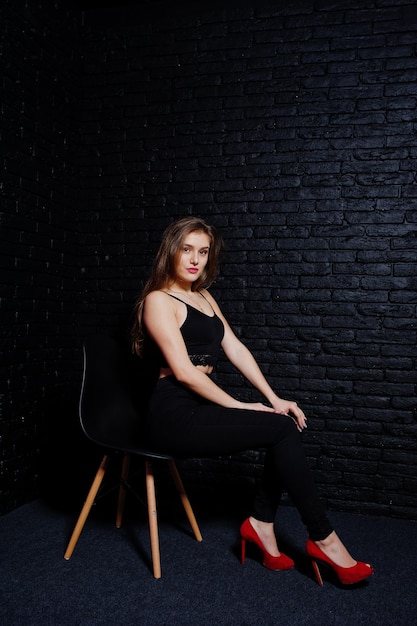 Hermosa chica morena vestida con tacones altos negros y rojos sentada y posando en una silla en el estudio contra la pared de ladrillo oscuro Retrato de modelo de estudio