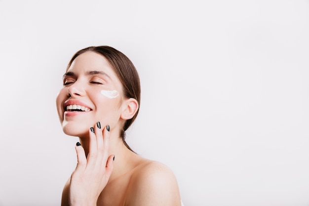 Hermosa chica con los ojos cerrados se ríe en la pared blanca. Modelo con crema sobre la piel que toca suavemente el rostro.