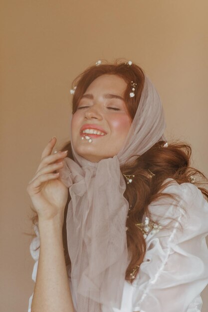 Hermosa chica con pañuelo ligero y blusa blanca se ríe con los ojos cerrados sobre fondo beige