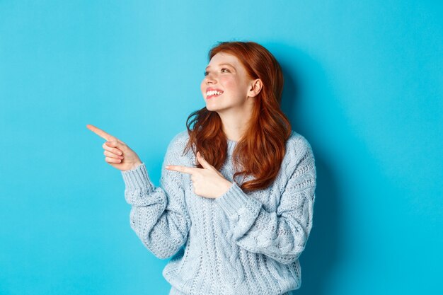 Hermosa chica pelirroja feliz, señalando con el dedo hacia la izquierda y mirando el logo complacido, de pie en suéter contra el fondo azul.