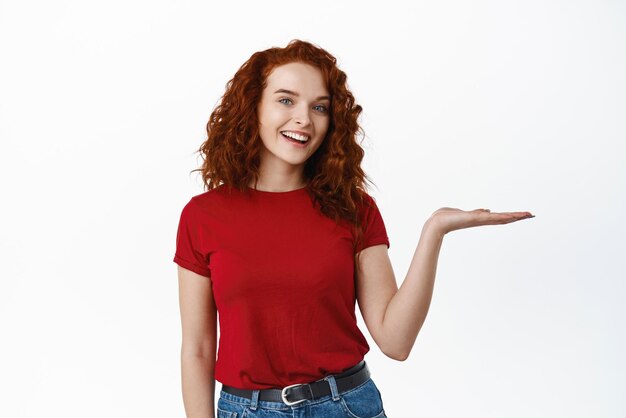 Hermosa chica pelirroja feliz sosteniendo en la mano sonriendo a la cámara y demostrando el producto en su palma de pie contra el fondo blanco