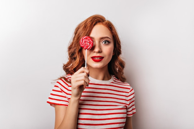 Hermosa chica pelirroja con piruleta posando en la pared blanca. Atractiva mujer joven con caramelo rojo.