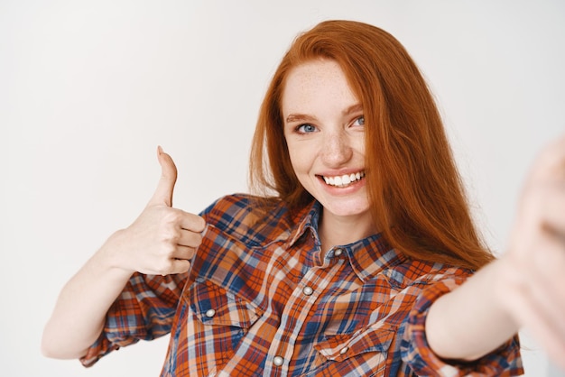 Foto gratuita hermosa chica pelirroja sonriendo feliz y mostrando el pulgar hacia arriba mientras toma selfie en la cámara frontal del teléfono inteligente ver fondo blanco