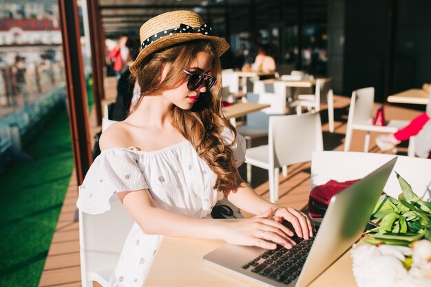 Hermosa chica con pelo largo con sombrero se sienta a la mesa en la terraza de la cafetería. Lleva un vestido blanco con hombros descubiertos, lápiz labial rojo, gafas de sol. Ella está escribiendo en la computadora portátil en serio.