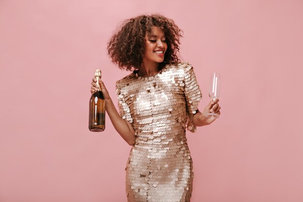 Hermosa chica de pelo mullido en vestido brillante de moda sonriendo con los ojos cerrados y sosteniendo un vaso y una botella con vino en la pared aislada.