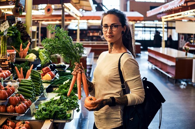 Hermosa chica pensativa con gafas está comprando zanahorias frescas en el mercado local de agricultores.