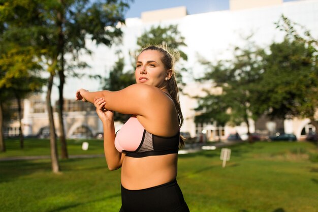 Hermosa chica pensativa de talla grande con top deportivo rosa y calzas mirando cuidadosamente a un lado mientras se estira en el parque de la ciudad