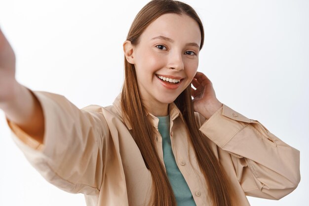 Foto gratuita hermosa chica posando para selfie, metiendo el pelo detrás de la oreja y sonriendo a la cámara frontal del teléfono inteligente, de pie contra el fondo blanco.