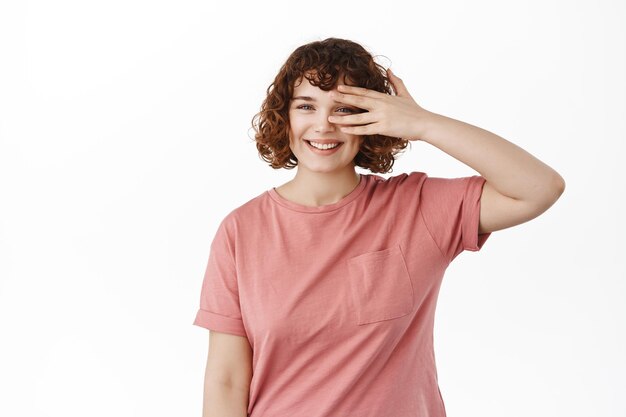 Hermosa chica rizada con cara pura y limpia, sin maquillaje, mirando a través de los dedos y sonriendo soñadora, con una camiseta rosa de verano, parada sobre fondo blanco