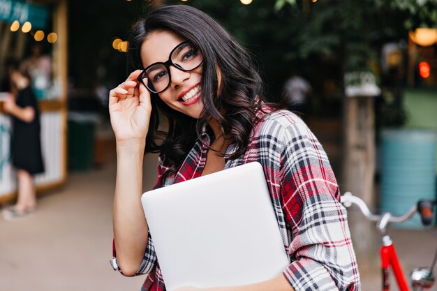 Hermosa chica rizada de pie en la calle con portátil. Foto al aire libre de estudiante inteligente en camisa a cuadros.