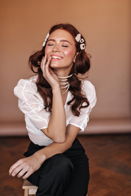 Hermosa chica rizada vestida con blusa blanca y pantalón negro se sienta en una silla Mujer con horquillas y collar de perlas posando en una habitación beige