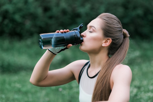 Hermosa chica en ropa deportiva, agua potable después del entrenamiento mientras está sentada en la hierba