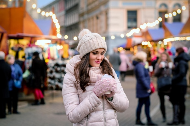 Hermosa chica con ropa de invierno se está calentando con una taza de café caliente parada en la calle con una feria durante la época navideña