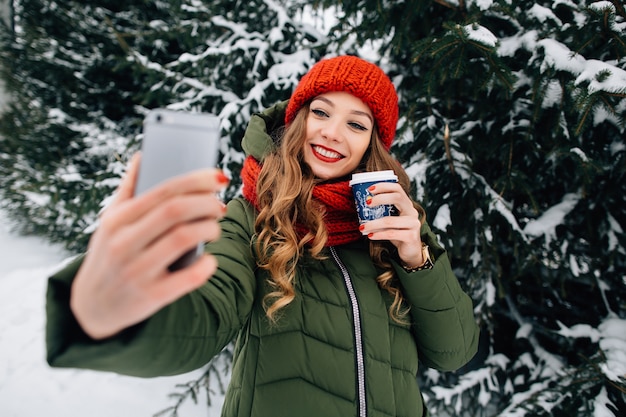 Hermosa chica en ropa de invierno toma selfie en smartphone. Mujer con café toma