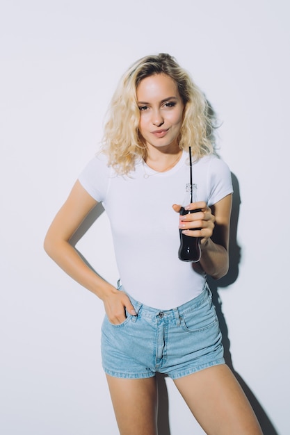 Hermosa chica rubia bebiendo cola de botella en ropa casual de verano en la pared blanca