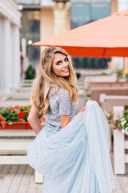 Hermosa chica rubia caminando sobre fondo de terraza. Ella sostiene una falda larga de tul azul en la mano y sonríe a la cámara.