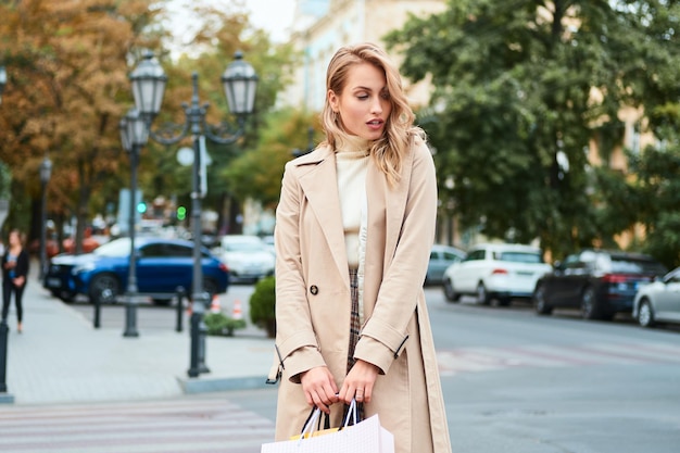 Hermosa chica rubia casual en elegante gabardina con bolsas de compras de pie en la calle de la ciudad