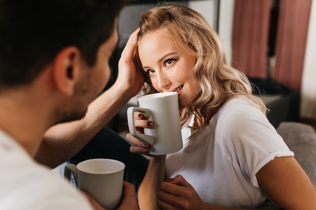 Hermosa chica rubia enamorada mirando a su novio y tomando café de la taza. Tierno lindo retrato de pareja romántica en casa.