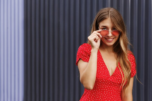 Hermosa chica rubia con gafas de sol rojas y vestido de verano sonriendo a la cámara, de pie al aire libre en un día soleado cerca de la pared de color